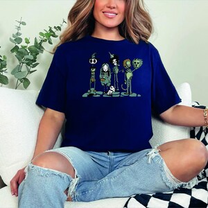 May include: A navy blue t-shirt with a graphic print of five cartoon characters. The characters are a skeleton, a witch, a vampire, a werewolf, and a zombie. The characters are all wearing costumes and are standing in a line. The t-shirt is a casual style with short sleeves and a crew neck.