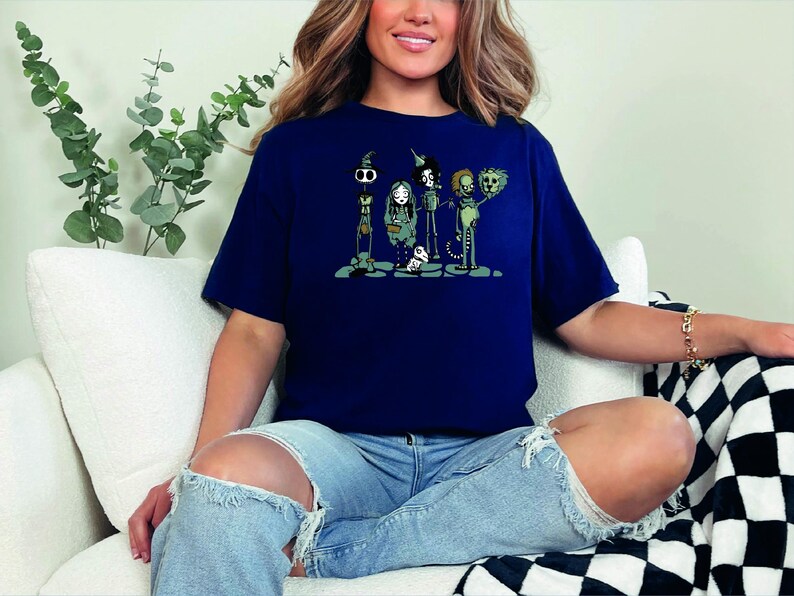 May include: A navy blue t-shirt with a graphic print of five cartoon characters. The characters are a skeleton, a witch, a vampire, a werewolf, and a zombie. The characters are all wearing costumes and are standing in a line. The t-shirt is a casual style with short sleeves and a crew neck.
