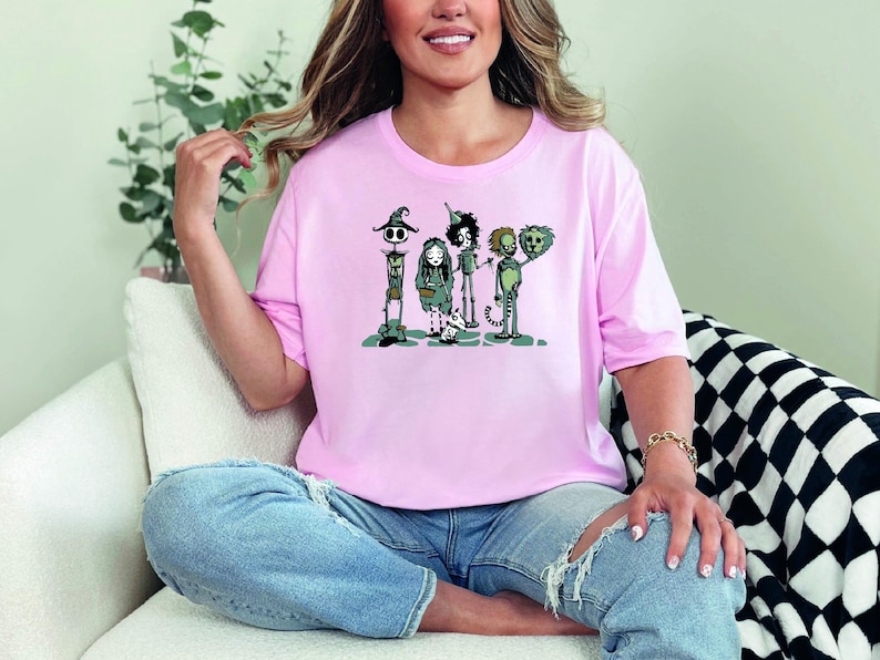 May include: A pink t-shirt with a graphic print of four cartoon characters. The characters are a skeleton, a witch, a tin man, and a lion. The characters are all wearing costumes and are standing in a line.