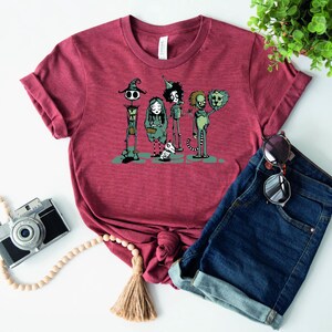May include: A maroon t-shirt with a graphic print of five cartoon characters. The characters are a witch, a girl, a boy, a creature with a crown, and a creature with a crown and a tail. The characters are all wearing different outfits and have different expressions.