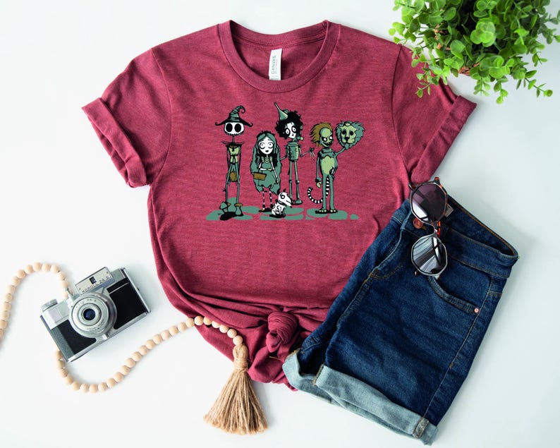 May include: A maroon t-shirt with a graphic print of five cartoon characters. The characters are a witch, a girl, a boy, a creature with a crown, and a creature with a crown and a tail. The characters are all wearing different outfits and have different expressions.