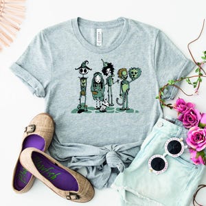 May include: A gray t-shirt with a graphic print of four cartoon characters. The characters are dressed in green and black and are standing in a line. The characters are from the movie "The Nightmare Before Christmas".