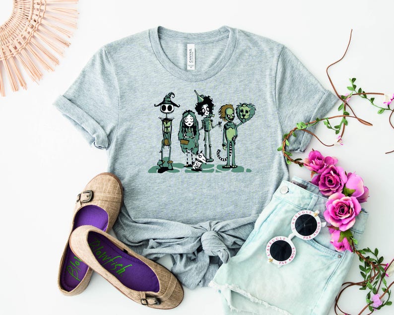 May include: A gray t-shirt with a graphic print of four cartoon characters. The characters are dressed in green and black and are standing in a line. The characters are from the movie "The Nightmare Before Christmas".