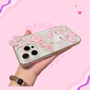 May include: A clear phone case with pink embellishments, including a bow, star, and Hello Kitty design. The case has a glittery background and a gold-colored frame. The phone case is held in a hand.