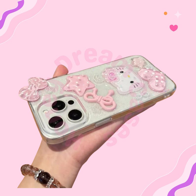 May include: A clear phone case with pink embellishments, including a bow, star, and Hello Kitty design. The case has a glittery background and a gold-colored frame. The phone case is held in a hand.