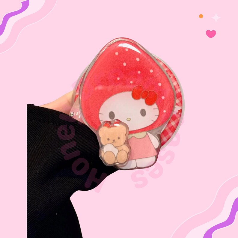 Puede incluir: Un espejo compacto transparente con forma de fresa, con un personaje de dibujos animados con un lazo y un vestido rojos, sosteniendo un peque&ntilde;o osito de peluche. El espejo tiene un dise&ntilde;o de lunares rojos y blancos y un borde a cuadros.
