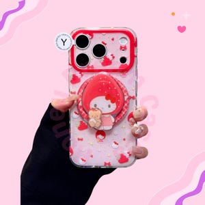 Puede incluir: Funda transparente para tel&eacute;fono con borde rojo y agarre emergente con un personaje de dibujos animados con un lazo rojo y un osito de peluche. La funda tiene un fondo rosa con patrones de fresas y lazos.