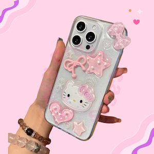 May include: Clear phone case decorated with pink bows, stars, and a heart. The case features a Hello Kitty design with a pink bow. The case is held in a hand against a pink background.