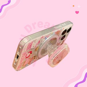 May include: Clear phone case with a pink pop-up grip featuring a Hello Kitty design. The case has a clear back with a pattern of Hello Kitty and other characters. The pop-up grip is pink and clear.
