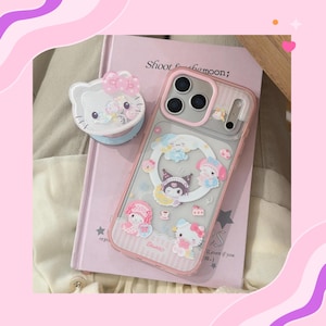 Op de afbeelding: Een roze telefoonhoesje met een doorzichtige achterkant en een roze rand, met stripfiguren. Een bijpassende Hello Kitty-telefoongreep ligt naast de telefoon. De hoes en de grip liggen op een roze boek met de tekst "Shoot for the moon;"