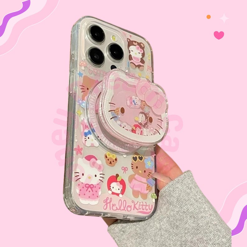May include: Clear phone case with Hello Kitty characters in pink and white. The case has a pop-up grip with a Hello Kitty design. The case is decorated with various Hello Kitty illustrations and the words "Hello Kitty".