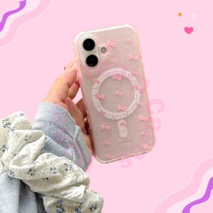 May include: Clear phone case with a pink Hello Kitty design. The case features a repeating pattern of pink bows and a circular magnetic charging area. The words "Hello Kitty" are printed at the bottom of the case.