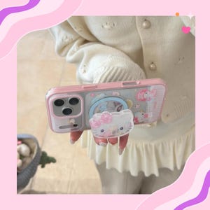 Op de afbeelding: Een roze telefoonhoesje met een doorzichtige achterkant en een pop-up grip met een Hello Kitty-ontwerp. De hoes heeft een transparant ontwerp met een roze rand en een pop-up grip met een Hello Kitty-ontwerp.