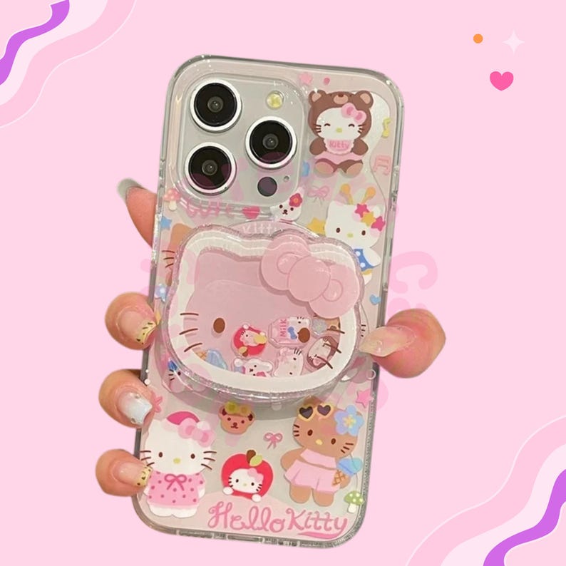 May include: Clear phone case with a pink Hello Kitty design and a pop-up grip. The case is decorated with various Hello Kitty characters and the text "Hello Kitty". The case is designed to fit a smartphone.