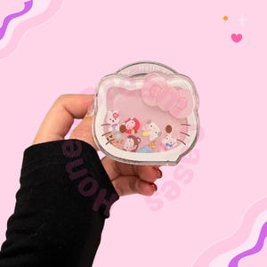 May include: A clear, pink, Hello Kitty-shaped case with a bow design. The case contains small, colourful character figures. The case is held against a pink background with decorative elements.