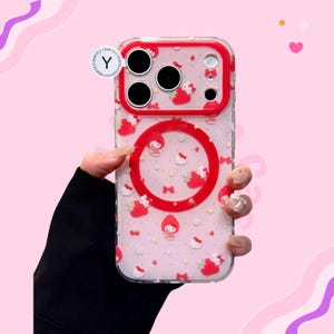 Puede incluir: Funda transparente para tel&eacute;fono con borde rojo y dise&ntilde;o circular rojo. La funda est&aacute; decorada con peque&ntilde;as figuras rojas de Hello Kitty, lazos y estrellas. La funda la sostiene una persona con una manga negra.