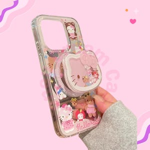 May include: Clear phone case with a pink Hello Kitty design and a pop-up grip. The case has a clear border and is decorated with various Hello Kitty characters and the text "Hello Kitty".