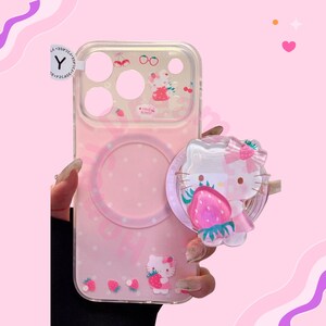 Puede incluir: Funda de tel&eacute;fono rosa con parte trasera transparente con dise&ntilde;o de Hello Kitty. La funda tiene un agarre emergente con un dise&ntilde;o de Hello Kitty y fresa. La funda tambi&eacute;n tiene un dise&ntilde;o de fresa y cereza.