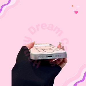 K&ouml;nnte beinhalten: Eine rosa Handyh&uuml;lle mit einem Cartoon-Charakter-Design. Die H&uuml;lle hat einen silbernen Rahmen und eine transparente R&uuml;ckseite mit einem hellrosa Design. Der Hintergrund ist rosa mit den Worten "Dream Case Shop".