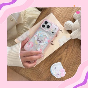 Op de afbeelding: Een doorzichtige telefoonhoes met een roze rand, met een cartoonontwerp met personages, wolken en cupcakes. De hoes wordt boven een boek op een houten oppervlak gehouden. Een bijpassende telefoonhouder is ook zichtbaar.