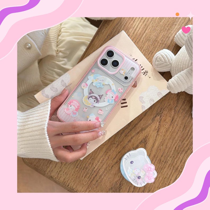 Op de afbeelding: Een doorzichtige telefoonhoes met een roze rand, met een cartoonontwerp met personages, wolken en cupcakes. De hoes wordt boven een boek op een houten oppervlak gehouden. Een bijpassende telefoonhouder is ook zichtbaar.