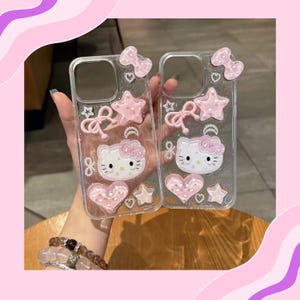 May include: Two clear phone cases decorated with pink bows, stars, hearts, and a Hello Kitty design. The cases have a glittery background and are held up by a person.