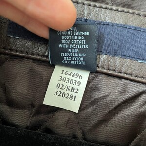 May include: Close-up of a brown leather jacket tag. The tag details the materials: genuine leather shell, acetate and polyester lining, and nylon and acetate sleeve lining. The tag also includes a series of numbers.