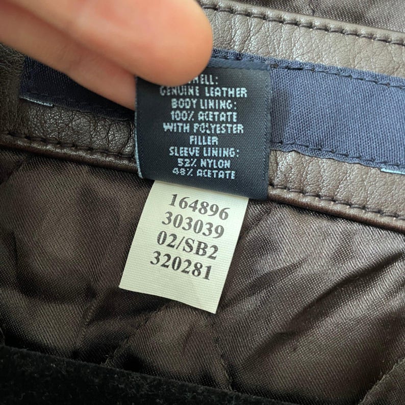 May include: Close-up of a brown leather jacket tag. The tag details the materials: genuine leather shell, acetate and polyester lining, and nylon and acetate sleeve lining. The tag also includes a series of numbers.