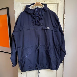 May include: A navy blue hooded pullover jacket featuring a partial button-up placket, a drawstring waist, and two front pockets with button closures. The chest displays the white embroidered HH Helly Hansen logo and text.