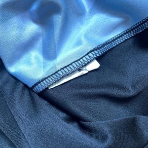 Puede incluir: Primer plano de una secci&oacute;n de ropa deportiva. La imagen muestra una tela azul claro y brillante cosida a una tela azul marino oscuro con hilo negro. Una etiqueta blanca con un logotipo es visible.