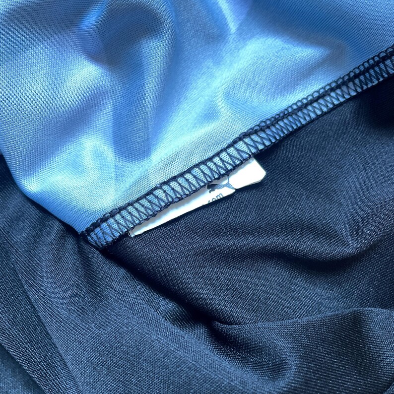 Puede incluir: Primer plano de una secci&oacute;n de ropa deportiva. La imagen muestra una tela azul claro y brillante cosida a una tela azul marino oscuro con hilo negro. Una etiqueta blanca con un logotipo es visible.