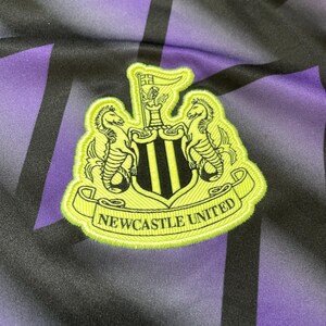 Puede incluir: Primer plano de un emblema del Newcastle United. El emblema est&aacute; bordado en verde ne&oacute;n y negro, con dos caballitos de mar que flanquean un escudo con tres rayas verticales negras. Las palabras "NEWCASTLE UNITED" est&aacute;n en una pancarta verde ne&oacute;n.
