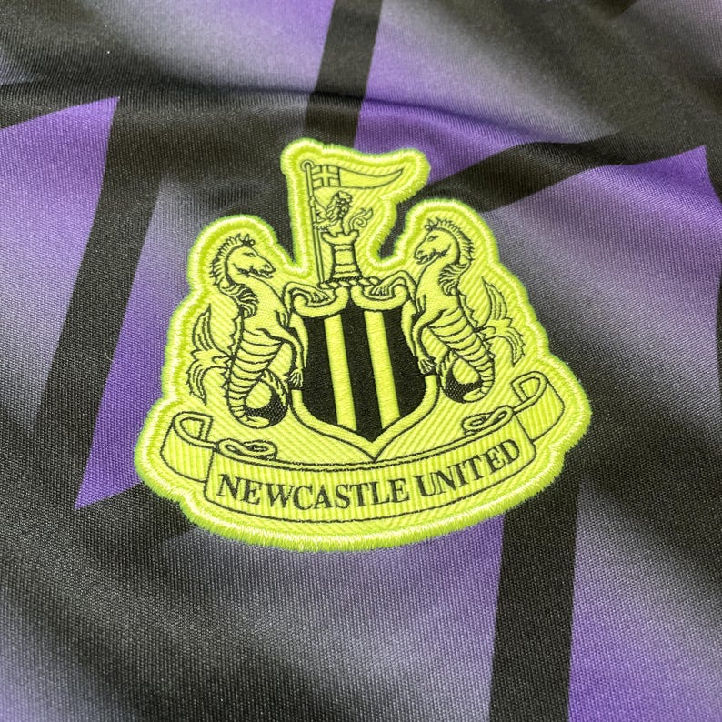 Puede incluir: Primer plano de un emblema del Newcastle United. El emblema est&aacute; bordado en verde ne&oacute;n y negro, con dos caballitos de mar que flanquean un escudo con tres rayas verticales negras. Las palabras "NEWCASTLE UNITED" est&aacute;n en una pancarta verde ne&oacute;n.