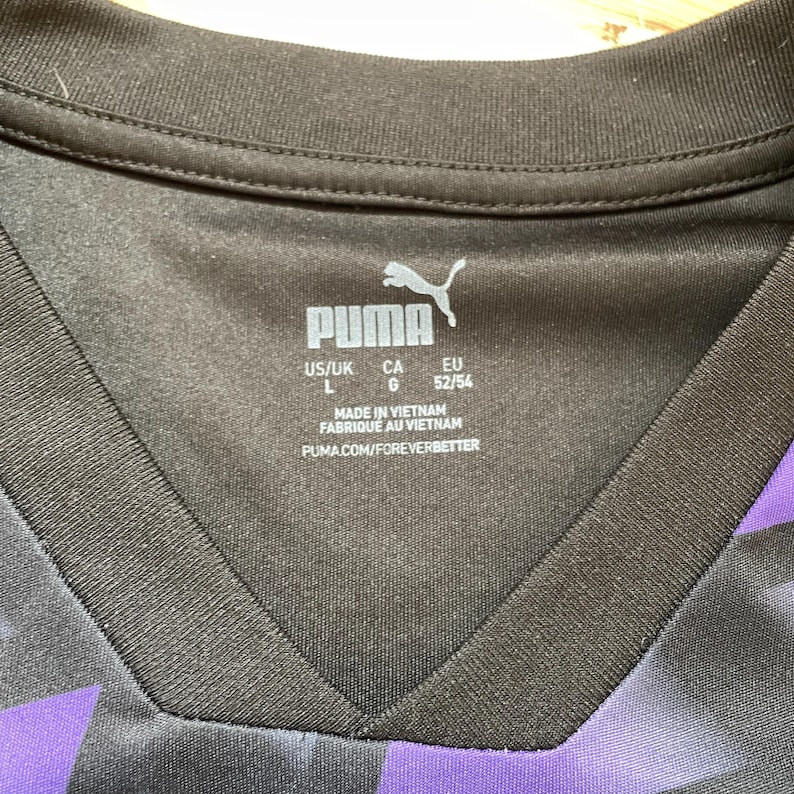 Puede incluir: Primer plano de una camiseta deportiva Puma negra y morada. El cuello de la camiseta presenta un dise&ntilde;o de cuello en V. El logotipo de Puma y la informaci&oacute;n de tallas son visibles. La camiseta est&aacute; hecha en Vietnam.