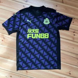 Puede incluir: Una camiseta de f&uacute;tbol negra y morada con un dise&ntilde;o geom&eacute;trico. La camiseta presenta el texto "FUN88" en verde ne&oacute;n, el logo de Puma y un escudo del equipo. Es de manga corta y tiene cuello en V.