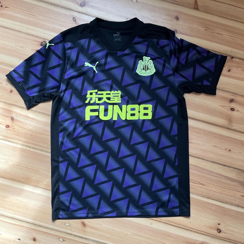 Puede incluir: Una camiseta de f&uacute;tbol negra y morada con un dise&ntilde;o geom&eacute;trico. La camiseta presenta el texto "FUN88" en verde ne&oacute;n, el logo de Puma y un escudo del equipo. Es de manga corta y tiene cuello en V.