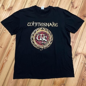 Può includere: T-shirt nera con il nome della band "Whitesnake" in lettere dorate sopra un emblema circolare. L'emblema ha un bordo dorato, dettagli rossi e dorati e le iniziali "WS".