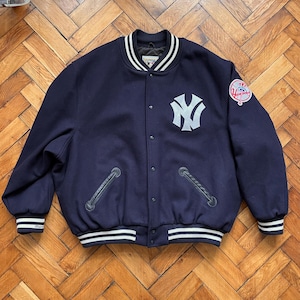 Puede incluir: Una chaqueta de béisbol de lana azul marino con ribetes de canalé blanco y negro. La chaqueta presenta un gran logotipo "NY" bordado en blanco en la parte delantera y un parche bordado más pequeño en la manga izquierda. La chaqueta tiene cierre de presión y dos bolsillos de cuero.