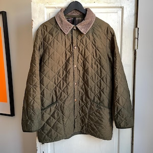 Könnte beinhalten: Olivgrüne Steppjacke mit Cordkragen und Knopfleiste. Die Jacke hat zwei Vordertaschen und hängt an einem schwarzen Kleiderbügel. Das Steppmuster ist ein klassisches Design.