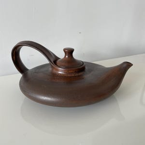 Può includere: Una teiera in ceramica marrone con un manico curvo e un piccolo coperchio. La teiera ha un corpo arrotondato e un beccuccio per versare. La teiera è di colore marrone pieno e sembra fatta a mano.
