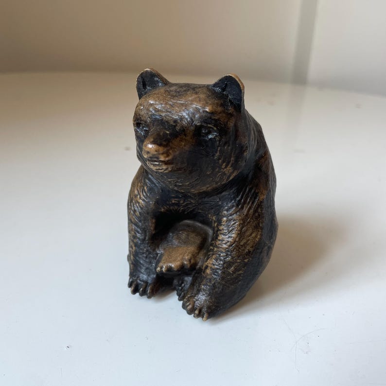 May include: A small, bronze-colored bear figurine in a seated position. The bear has a detailed texture, with visible fur and a focused expression. The figurine is a decorative item, likely for display.