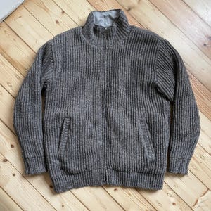 May include: A gray, ribbed-knit zip-up sweater with a high collar and two front pockets. The sweater is made of a thick, textured material and is laid flat on a wooden surface. The zipper runs down the center of the sweater.