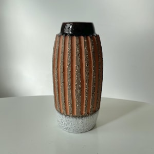May include: A ceramic vase with a textured, ribbed design. The vase has vertical stripes in shades of brown and orange, with a speckled texture. The top is dark brown, and the base is white with a speckled pattern. The vase is a decorative item.