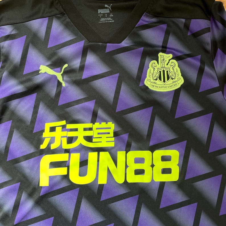Puede incluir: Camiseta de f&uacute;tbol negra y morada con un patr&oacute;n geom&eacute;trico. La camiseta presenta el logo de Puma en verde lima, junto con el texto "乐天堂 FUN88" en amarillo. Tambi&eacute;n es visible el escudo del Newcastle United.