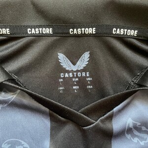 May include: Black and gray athletic jersey with the brand name "CASTORE" printed on the collar. The jersey features a V-neck design and a logo with the brand name and size labels.