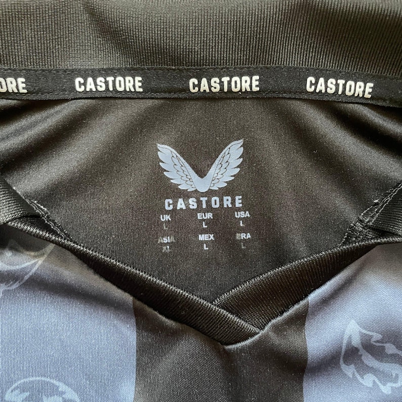 May include: Black and gray athletic jersey with the brand name "CASTORE" printed on the collar. The jersey features a V-neck design and a logo with the brand name and size labels.