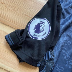 May include: Close-up of a black and blue soccer jersey sleeve. The sleeve features a Premier League logo and a date, 09/12/1892, printed on the jersey. The jersey has a patterned design.