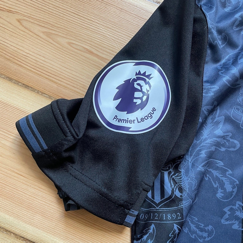 May include: Close-up of a black and blue soccer jersey sleeve. The sleeve features a Premier League logo and a date, 09/12/1892, printed on the jersey. The jersey has a patterned design.