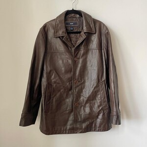 May include: A brown leather jacket with a button-up front and a collared neckline. The jacket has two front pockets and is hanging on a black hanger. The brand name "GAP" is visible on the label.