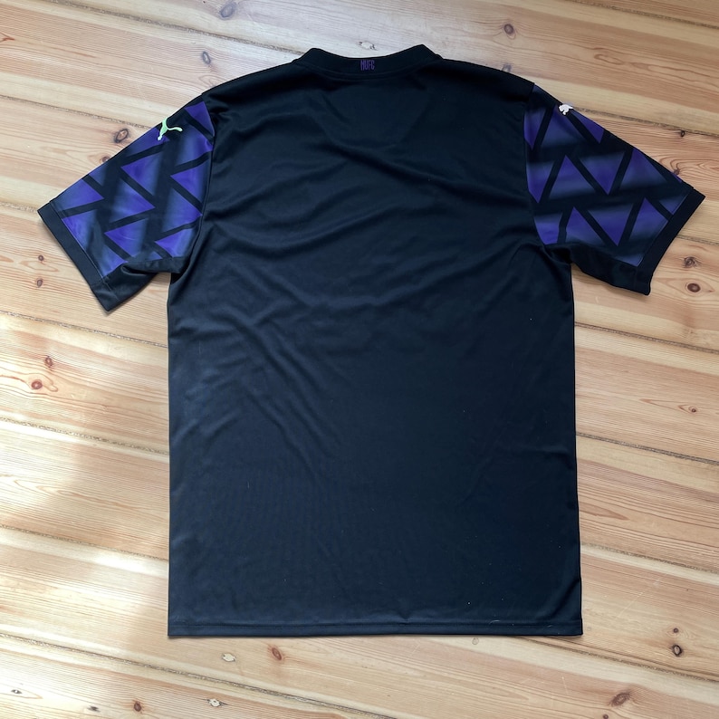 Puede incluir: Camiseta deportiva negra con mangas estampadas geom&eacute;tricas moradas y azules. La camiseta tiene cuello en V y un peque&ntilde;o logotipo en la parte superior de la espalda. Las mangas tienen un dise&ntilde;o de tri&aacute;ngulo repetido. La camiseta est&aacute; hecha de un material ligero y transpirable.